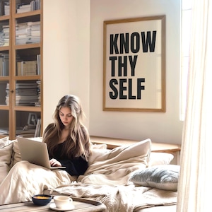 May include: A woman sits on a couch with a laptop in her lap. The couch is covered in a beige blanket and there is a wooden coffee table in front of her. A framed print with the text "KNOW THY SELF" hangs on the wall behind her.