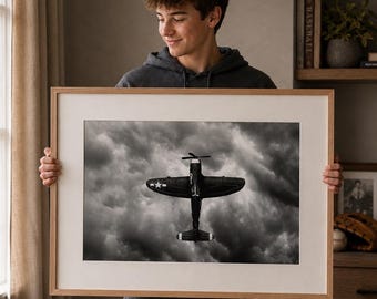 May include: A framed wall art print featuring a black and white photograph of a vintage propeller airplane flying through dark, dramatic clouds. The artwork is encased in a light-colored wooden frame with a wide white mat border surrounding the image.