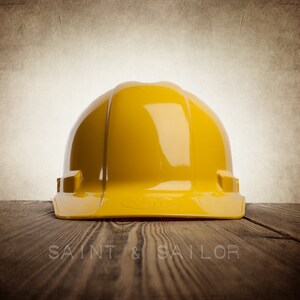 May include: A bright yellow hard hat, a safety helmet, sits on a wooden surface. The hard hat is the primary focus, with a glossy finish. The background is a textured, sepia-toned wall. The words "SAINT & SAILOR" are at the bottom.