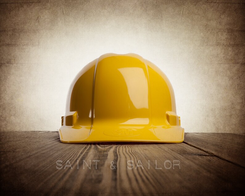 May include: A bright yellow hard hat, a safety helmet, sits on a wooden surface. The hard hat is the primary focus, with a glossy finish. The background is a textured, sepia-toned wall. The words "SAINT & SAILOR" are at the bottom.