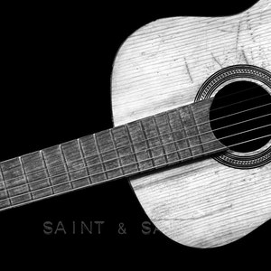 May include: A black and white photo of an acoustic guitar with a worn wooden body and a soundhole. The guitar is lying on a black surface. The text "SAINT & SA" is visible in the lower left corner of the image.