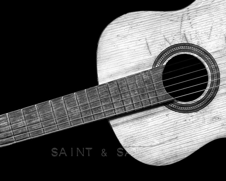 May include: A black and white photo of an acoustic guitar with a worn wooden body and a soundhole. The guitar is lying on a black surface. The text "SAINT & SA" is visible in the lower left corner of the image.