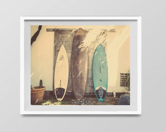 May include: Three surfboards leaning against a weathered wooden door. The surfboards are different colours and styles, with one being white, one being brown, and one being blue. The surfboards are in front of a white wall with a wooden door and a window.