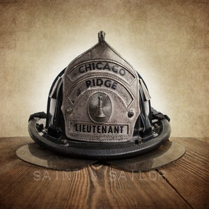 May include: A vintage firefighter helmet with a silver badge that reads "Chicago Ridge Lieutenant". The helmet is black with a silver trim and is sitting on a wooden surface.