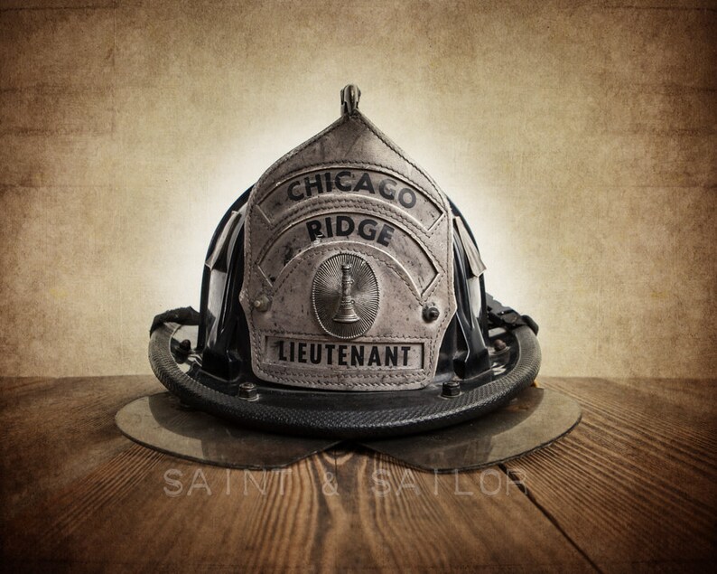 May include: A vintage firefighter helmet with a silver badge that reads "Chicago Ridge Lieutenant". The helmet is black with a silver trim and is sitting on a wooden surface.