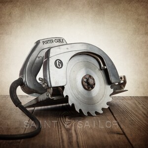 May include: Vintage Porter-Cable circular saw with a silver metal housing and a large, circular blade. The saw has a black power cord and is resting on a wooden surface. The number "6" is visible on the side of the saw.