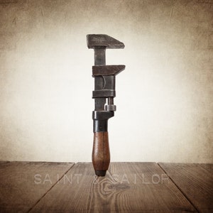May include: An antique adjustable wrench with a dark metal head and a wooden handle. The wrench is positioned vertically against a neutral background, with the text "SAINT & SAILOR" visible at the bottom. The tool rests on a wooden surface.