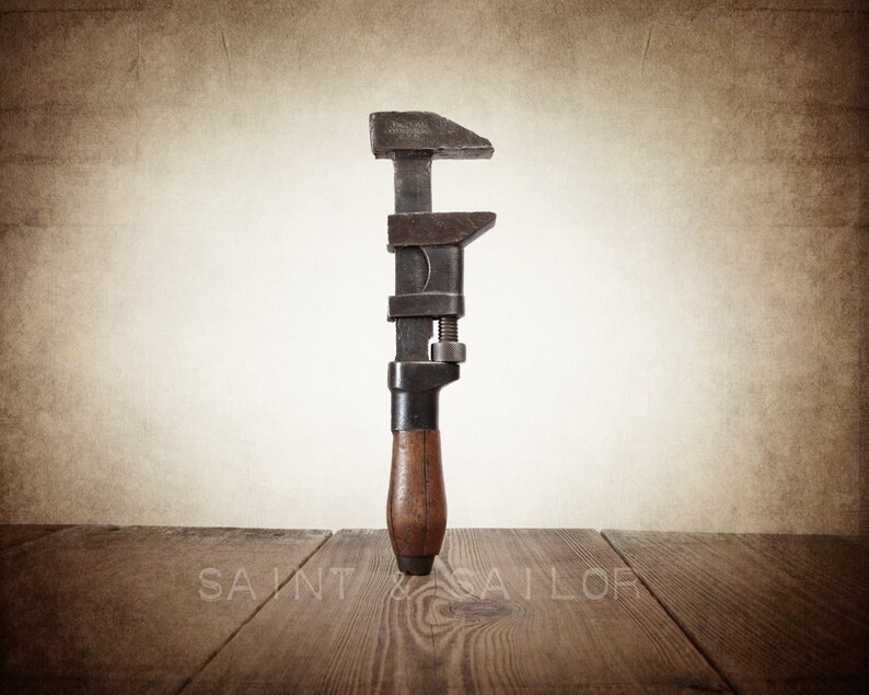 May include: An antique adjustable wrench with a dark metal head and a wooden handle. The wrench is positioned vertically against a neutral background, with the text "SAINT & SAILOR" visible at the bottom. The tool rests on a wooden surface.
