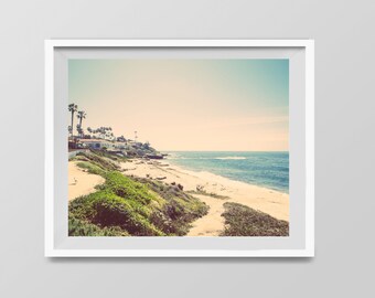 May include: A vintage-style photograph of a beach scene with a sandy shore, blue ocean, and a line of palm trees and houses in the distance.