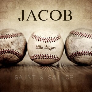 May include: Three vintage baseballs with red stitching on a wooden surface. The middle baseball has the text "little slugger" written on it. The text "JACOB" is at the top of the image and the text "SAINT & SAILOR" is at the bottom of the image.