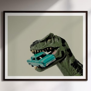 Op de afbeelding: Een ingelijste kunstdruk met een groen Tyrannosaurus Rex-speeltje met donkere vlekken, dat een kleine, talingkleurige vintage speelgoedauto in zijn open kaken houdt. De dinosaurus heeft een getextureerde huid en zichtbare tanden.