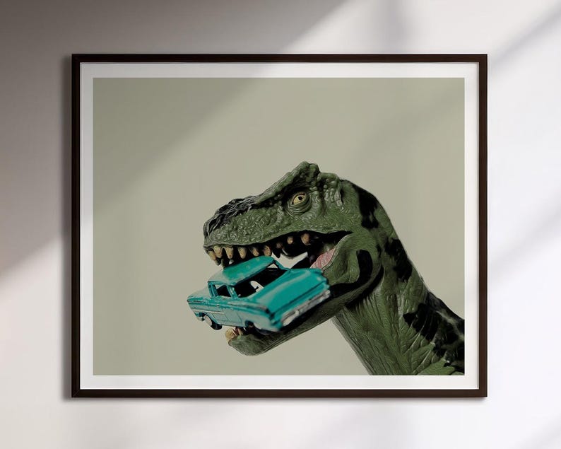Op de afbeelding: Een ingelijste kunstdruk met een groen Tyrannosaurus Rex-speeltje met donkere vlekken, dat een kleine, talingkleurige vintage speelgoedauto in zijn open kaken houdt. De dinosaurus heeft een getextureerde huid en zichtbare tanden.