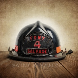 May include: A black firefighter helmet with a red FDNY 4 SALVAGE patch. The helmet is sitting on a wooden surface.