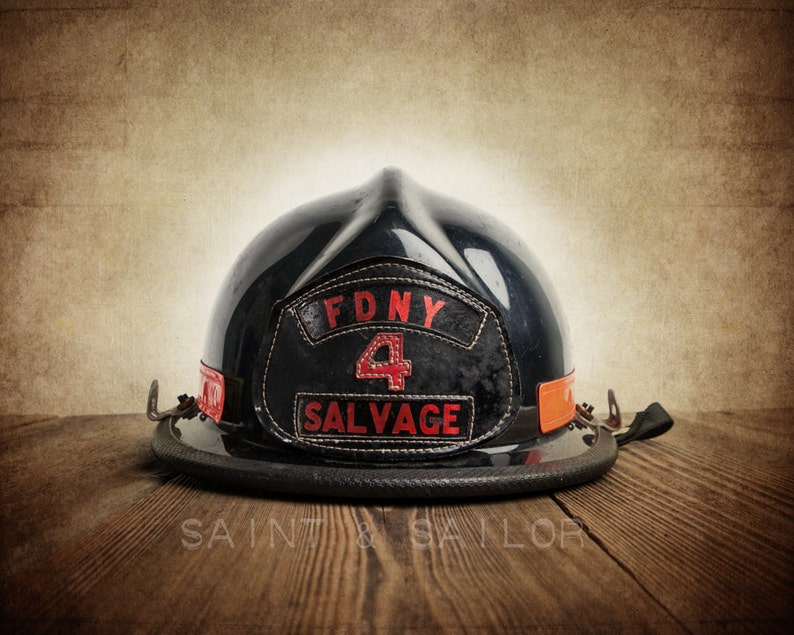 May include: A black firefighter helmet with a red FDNY 4 SALVAGE patch. The helmet is sitting on a wooden surface.
