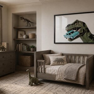Op de afbeelding: Een ingelijste kunstdruk met een groene Tyrannosaurus Rex die in een lichtblauwe vintage auto bijt. Het kunstwerk hangt in een kamer met een houten peuterbed, een dressoir, planken met dinosaurusdecoratie en een getextureerd vloerkleed.
