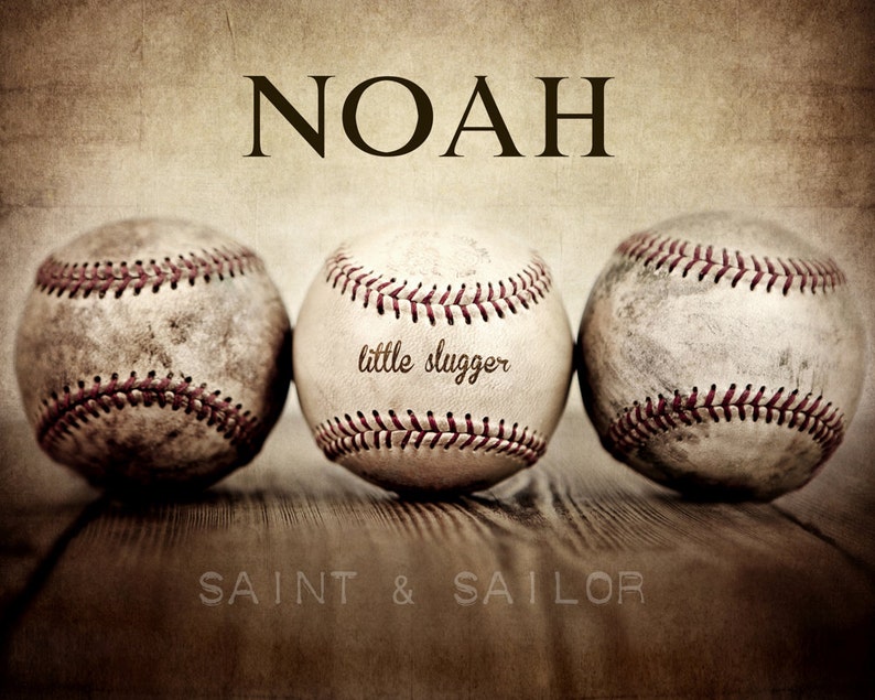 May include: Three vintage baseballs with red stitching, one with the text "little slugger" on it, arranged on a wooden surface with the name "NOAH" above and the text "SAINT & SAILOR" below.