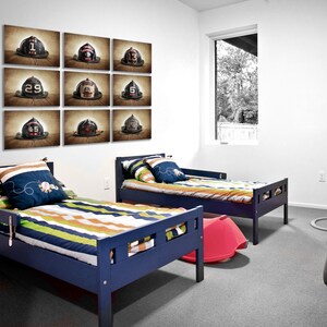 May include: Two blue twin beds with striped bedding in a room with a white wall and a window. Nine framed pictures of vintage fire helmets are displayed on the wall above the beds.