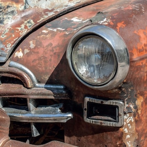 May include: Close-up of a vintage car's front end, showcasing a round headlight with a chrome bezel. The car's body exhibits extensive rust and peeling paint in shades of orange, brown, and black, suggesting age and weathering. The grill is also visible.