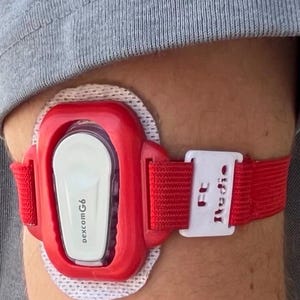 Puede incluir: Una funda protectora de plástico rojo para un monitor continuo de glucosa Dexcom G6, con un inserto central blanco y una correa elástica roja con un clip de ajuste de plástico blanco etiquetado con el texto FC Studio, asegurada sobre un parche adhesivo blanco.