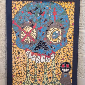 Puede incluir: Una pieza de arte mural de mosaico rectangular en un marco negro que presenta una calavera azul estilizada con una cruz roja sobre un ojo, dientes blancos y detalles verdes sobre un fondo amarillo, con un pequeño personaje circular abajo a la derecha.