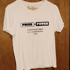 Può includere: Maglietta bianca con la scritta nera "PRIDE = POWER" e la scritta "CHRISTOPHER STREET GAY LIBERATION DAY 1970". La maglietta è appesa a una gruccia di legno su uno sfondo di legno.