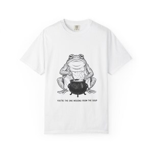May include: White t-shirt featuring a black and white illustration of a frog wearing an apron and stirring a pot. The text "YOU'RE THE ONE MISSING FROM THE SOUP" is printed below the frog.