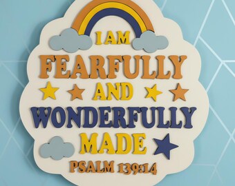 May include: A cloud-shaped wall sign with a cream background featuring a rainbow, stars, and clouds. The text reads: I AM FEARFULLY AND WONDERFULLY MADE PSALM 139:14. The lettering is in orange, yellow, and dark blue, with layered 3D wooden cutouts.