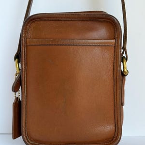 May include: A rectangular brown leather crossbody bag featuring a slim exterior slip pocket, a long adjustable shoulder strap, and brass hardware. A leather hangtag is attached to the side by a small brass ball chain.