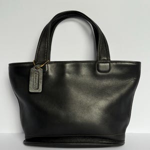 May include: A black leather handbag featuring two top handles and a rectangular leather hang tag embossed with the word COACH. The bag has a smooth finish, a wide opening, and a structured base with visible stitching along the bottom edge.