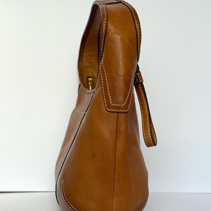 May include: A tan leather shoulder bag with a curved design. The bag features a single handle and a small strap. White stitching accents the seams. The bag is set against a white background.
