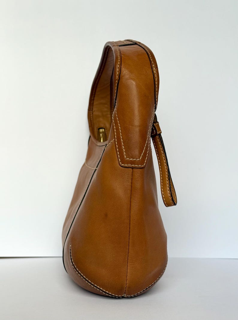 May include: A tan leather shoulder bag with a curved design. The bag features a single handle and a small strap. White stitching accents the seams. The bag is set against a white background.