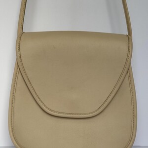 May include: A beige leather crossbody bag with a curved flap. The bag features a long, thin strap and is made of smooth leather. Stitching is visible around the edges of the bag.