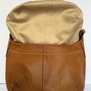 May include: A tan leather handbag with a gold satin lining. The bag features white stitching and a curved design. The bag is open, showing the interior. The leather has a natural, slightly worn look.