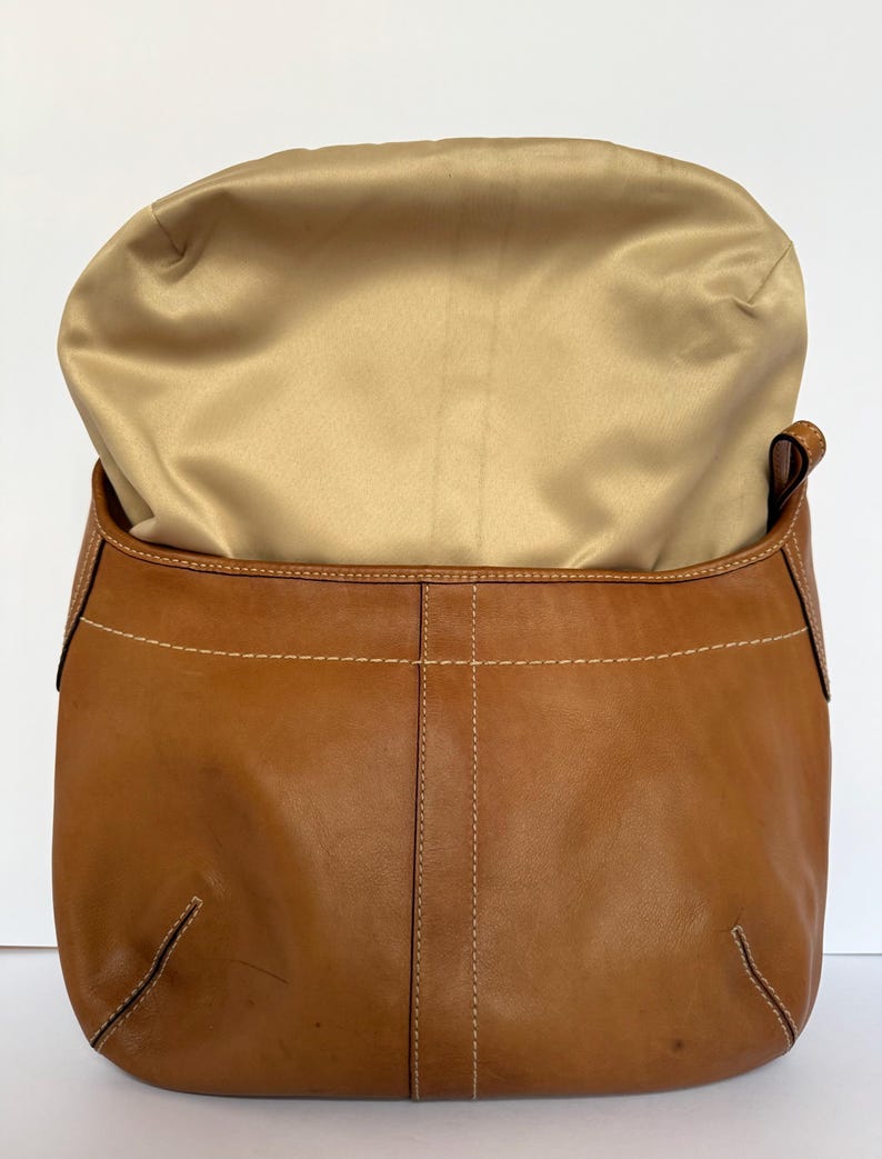 May include: A tan leather handbag with a gold satin lining. The bag features white stitching and a curved design. The bag is open, showing the interior. The leather has a natural, slightly worn look.