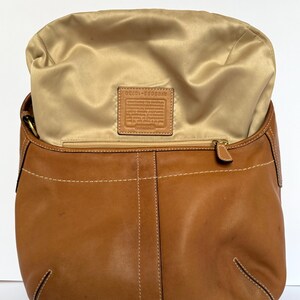 May include: A tan leather handbag with a gold satin interior. The bag has a curved shape with white stitching details and a zip closure. A small leather tag is visible inside the bag.