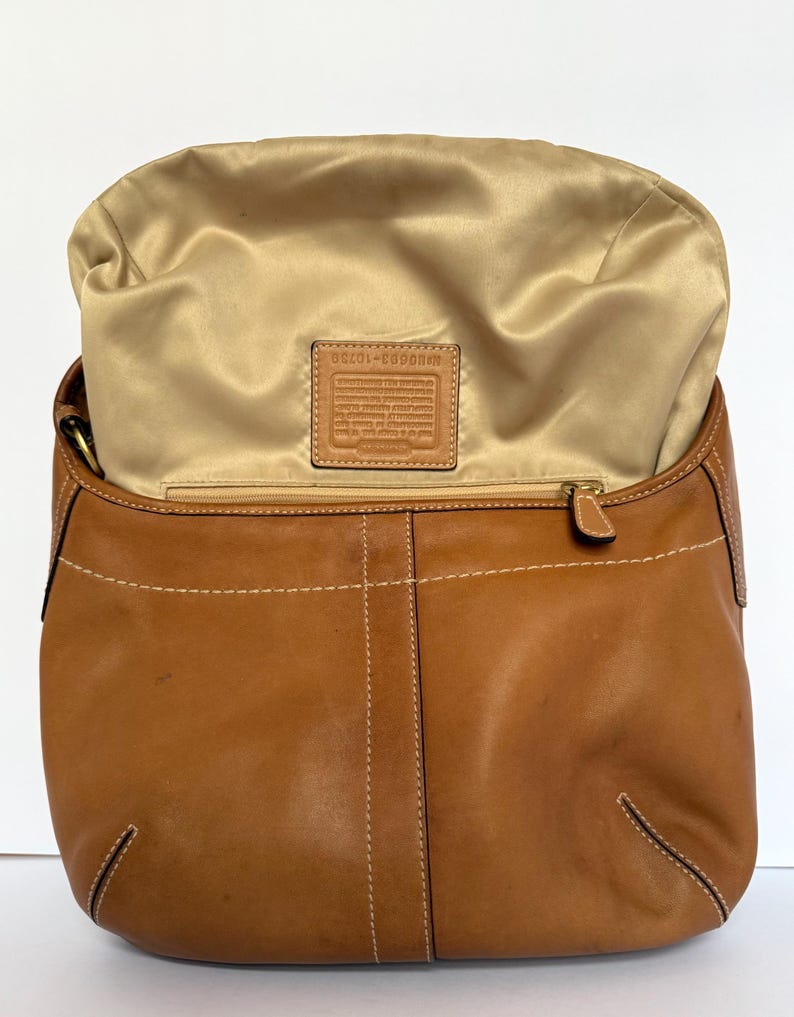 May include: A tan leather handbag with a gold satin interior. The bag has a curved shape with white stitching details and a zip closure. A small leather tag is visible inside the bag.