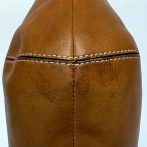 May include: Close-up of a tan leather bag. The bag has a rounded shape with a stitched seam down the centre and white stitching along the top. The leather has a slightly distressed appearance.