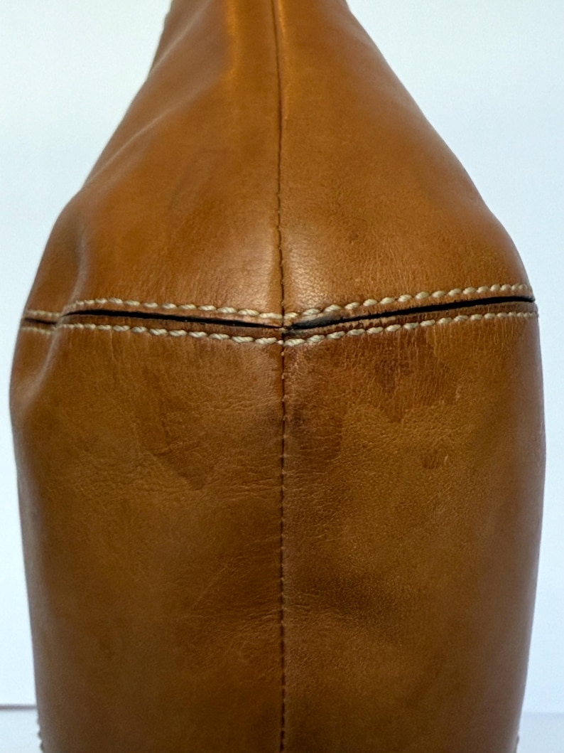 May include: Close-up of a tan leather bag. The bag has a rounded shape with a stitched seam down the centre and white stitching along the top. The leather has a slightly distressed appearance.