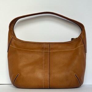 May include: A tan leather shoulder bag with a curved handle. The bag features visible stitching in a lighter colour, and a zip closure. The bag is a medium size and has a smooth, slightly worn appearance.