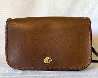 May include: A brown leather crossbody bag with a rectangular shape and rounded edges. The bag features a gold-tone clasp and a long, adjustable strap. The leather has a slightly worn appearance.