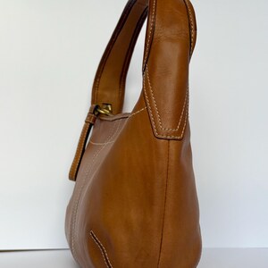 May include: A brown leather shoulder bag with a curved shape. The bag has a single handle and a gold-coloured buckle. White stitching accents the seams. The bag is made of smooth leather.