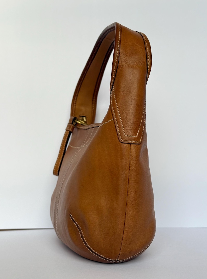May include: A brown leather shoulder bag with a curved shape. The bag has a single handle and a gold-coloured buckle. White stitching accents the seams. The bag is made of smooth leather.