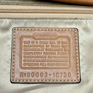 May include: Close-up of a Coach bag tag. The tag is rectangular with a brown leather appearance and black stitching. Text on the tag reads: "THIS IS A COACH BAG. IT WAS HANDCRAFTED IN CHINA..." and "N&deg; M0693-10739."