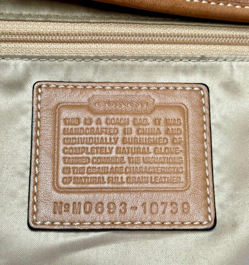 May include: Close-up of a Coach bag tag. The tag is rectangular with a brown leather appearance and black stitching. Text on the tag reads: "THIS IS A COACH BAG. IT WAS HANDCRAFTED IN CHINA..." and "N&deg; M0693-10739."