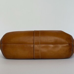 May include: A tan leather handbag with white stitching. The bag has a rounded shape and a central vertical seam. The stitching is visible along the edges and the central seam. The bag is empty and sits on a white surface.
