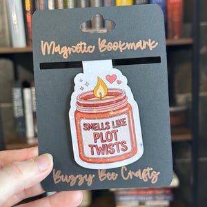 May include: A black magnetic bookmark with the words "Magnetic Bookmark" in copper-colored script. The bookmark features a sticker of a candle jar with the text "Smells like Plot Twists". The brand name "Buzzy Bee Craftz" is also visible.