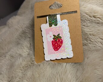 May include: A strawberry-themed sticker on a brown card. The sticker has a pink background with a white flower and a red strawberry. The sticker has a white, glittery border. The card has a hole at the top for hanging.