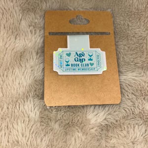 May include: A brown card with a die-cut hole at the top, holding a teal and iridescent bookmark. The bookmark reads "Age Gap Book Club Lifetime Membership" with decorative hearts and hourglasses.