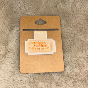 May include: A brown cardboard tag with a die-cut hole at the top, holding a rectangular sticker. The sticker reads "Grumpy Sunshine Book Club Lifetime Membership" in orange and white, with heart and sun graphics.