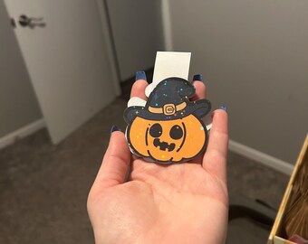 Könnte beinhalten: Ein Halloween-Aufkleber in Kürbisform mit Hexenhut. Der Kürbis ist orange mit einem lächelnden Gesicht und Glitzerakzenten. Der Hexenhut ist schwarz mit einer goldenen Schnalle und Glitzersternen.